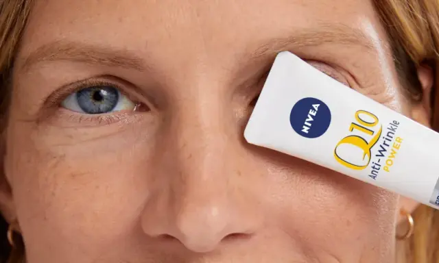 Un tube de crème pour les yeux Nivea Q10 Anti-Wrinkle Power est tenu devant l'œil d'une femme, masquant les rides pattes d'oie.