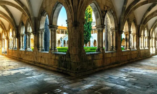 Gotische Arkaden im Kloster Batalha, Portugal. Sonnenschein fällt auf den Steinboden und beleuchtet den grünen Innenhof.