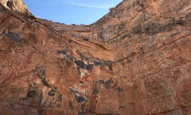 Acantilado de roca rojiza con vetas oscuras, mostrando diferentes tipos de canteras y la estratificación geológica bajo un cielo azul.