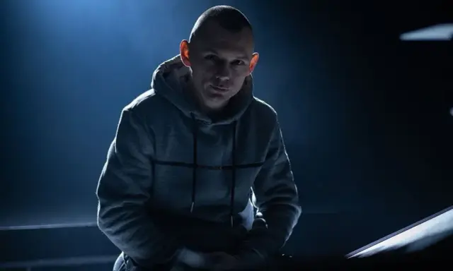 Młody mężczyzna w szarej bluzie z kapturem, z ogoloną głową, wygląda jak jeden z popularnych raperów, gotowy do występu.