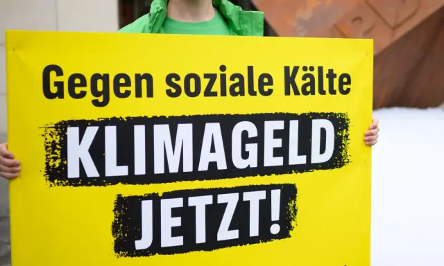 Gelbe Tafel mit schwarzer Schrift: "Gegen soziale Kälte KLIMAGELD JETZT!". Eine Forderung nach sozialer Gerechtigkeit im Klimaschutz.