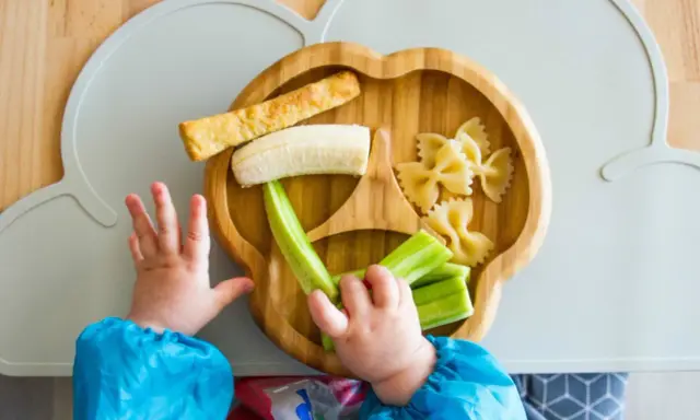 Babyhände greifen nach leckeren Baby-Snacks ab 6 Monaten: Banane, Gurke, Nudeln und ein Stück Brot auf einem Holztablett.