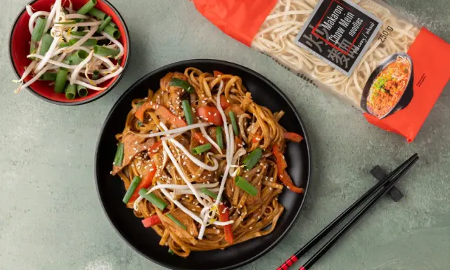 Pyszny makaron chow mein z warzywami i kiełkami fasoli na czarnym talerzu, obok paczka makaronu i miseczka kiełków.