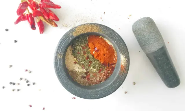 Mortier en pierre rempli d'épices pour fajitas, avec des piments rouges séchés et des grains de poivre à proximité.