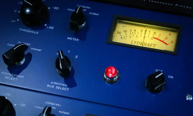 Gros plan sur un compresseur audio bleu avec des boutons, un VU-mètre et une lumière rouge allumée.
