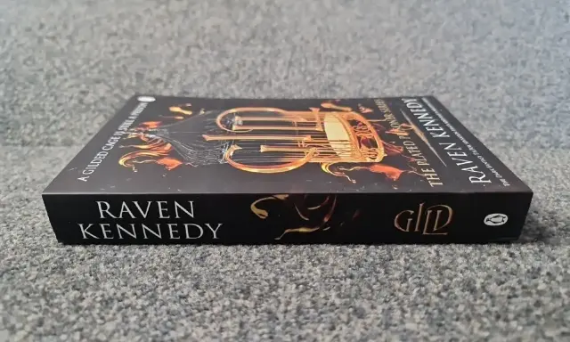 Okładka książki "A Gilded Cage is Still a Prison" autorstwa Raven Kennedy. Złota klatka i płomienie na ciemnym tle.
