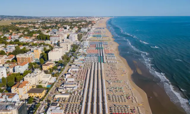 Vista aerea delle frazioni di Rimini: una lunga spiaggia attrezzata con file ordinate di ombrelloni e lettini, hotel e palazzi affacciati sul mare Adriatico.