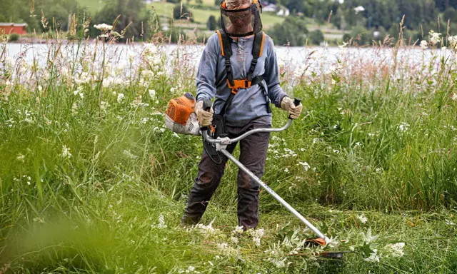 Persona con equipo de protección usa desbrozadora para cortar hierba alta, mostrando la diferencia entre desbrozadora y cortabordes en un paisaje rural.