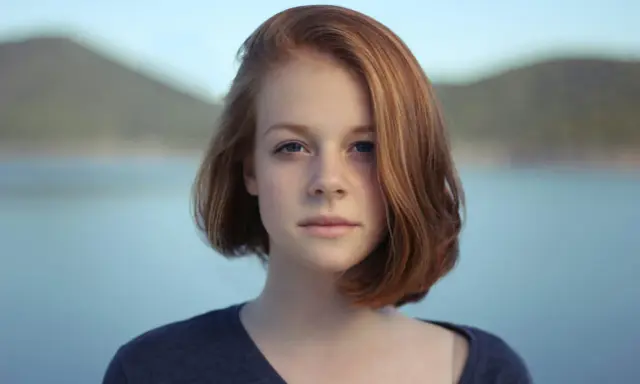 Ritratto fotografia di una giovane ragazza con capelli rossi e occhi azzurri, sullo sfondo di un lago e colline.