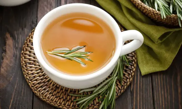 Une tasse d'infusion romarin dorée, garnie d'une branche de romarin frais, sur un dessous de plat en osier.