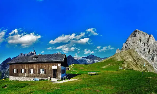 Rifugio Passo San Nicolò, un'accogliente baita in legno con vista su maestose cime innevate e un cielo azzurro brillante.
