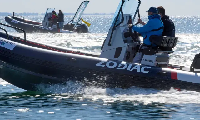 Deux bateaux Zodiac naviguent sur l'eau. L'un d'eux, au premier plan, porte le logo "ZODIAC" et semble communiquer sur le canal 9 VHF.