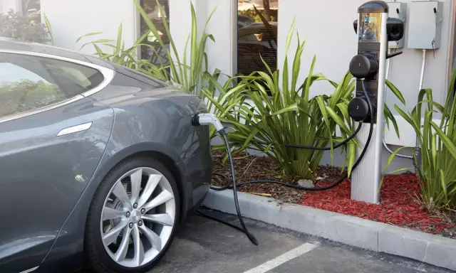 Coche eléctrico gris cargando en uno de los cargadores públicos. La estación tiene dos puntos de carga y está rodeada de vegetación.