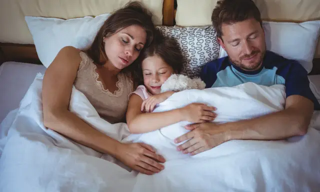 Familia durmiendo junta, la niña abraza un peluche. Un momento de paz que evoca la regresión infantil y la seguridad del hogar.