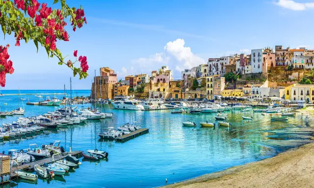 Un pittoresco porto con barche ormeggiate, case colorate su una collina e fiori rossi in primo piano, un'immagine che evoca la bellezza di Sardegna e Sicilia.