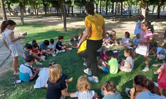 Niños sentados en el césped, participando en una gymkana para niños. Un adulto con camiseta amarilla dirige la actividad.