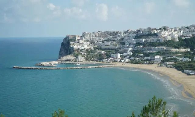 En vit stad klättrar uppför en klippa vid havet i Puglia, Italien. En strand och en hamn syns nedanför.