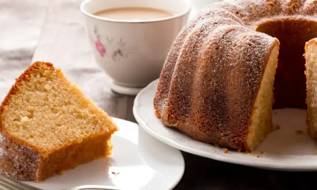 Una fetta di torta al latte soffice e dorata, cosparsa di zucchero a velo, accanto a una ciambella intera e una tazza di tè.