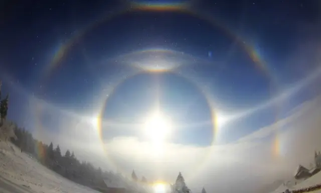 Zjawiskowy efekt halo nad zimowym krajobrazem. Słońce otoczone tęczowymi kręgami, mgła i ośnieżone drzewa tworzą magiczną scenę.