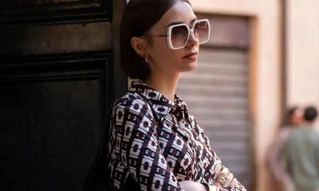Emily in Paris, Lily Collins poses with large white sunglasses and a patterned shirt, leaning against a dark door.