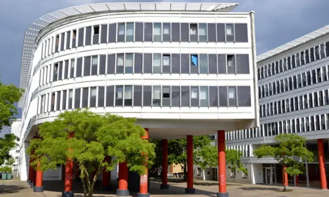 Edificio curvo blanco con ventanas oscuras y árboles verdes. Columnas rojas sostienen la estructura, creando un espacio crous.