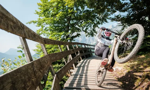 Ciclista in azione durante il giro del lago di Molveno in bici, su un ponte di legno tra alberi verdi e montagne.