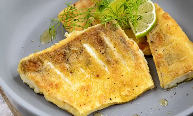Goldbraunes Zanderfilet, knusprig gebraten, serviert mit Limettenspalten und Dill auf einem grauen Teller.