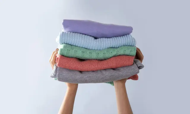 Manos sostienen una pila de ropa doblada en tonos pastel. Consejos para saber como evitar que una prenda destiña.