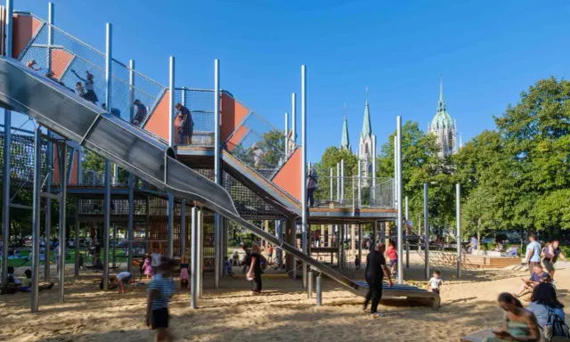 Un día perfecto de **munich con niños** en un parque infantil moderno con toboganes y estructuras de juego, con torres de iglesias al fondo.