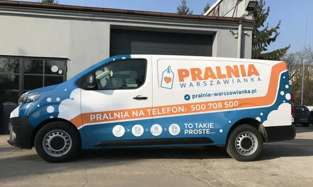 Biało-niebieski van z pomarańczowymi elementami to reklama tranzytowa dla pralni. Na boku widnieje napis "PRALNIA WARSZOWIANKA" i numer telefonu.