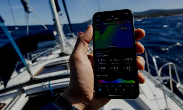 Sur un voilier, une main tient un smartphone affichant une application météo marine gratuite avec les prévisions pour Ibiza.