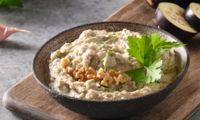 En skål med krämig baba ganoush, toppad med valnötter och persilja. Bredvid ligger skivade auberginer och ett vitlöksklyfta.