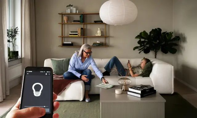 En kvinna och en tonåring kopplar av i ett vardagsrum med modern, begagnad inredning. En mobiltelefon visar en ljusapp.