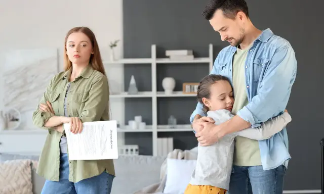 Ojciec tuli córkę, podczas gdy matka z dokumentem rozwodowym patrzy z boku. Przejęcie opieki nad dzieckiem przez ojca w trudnym momencie.