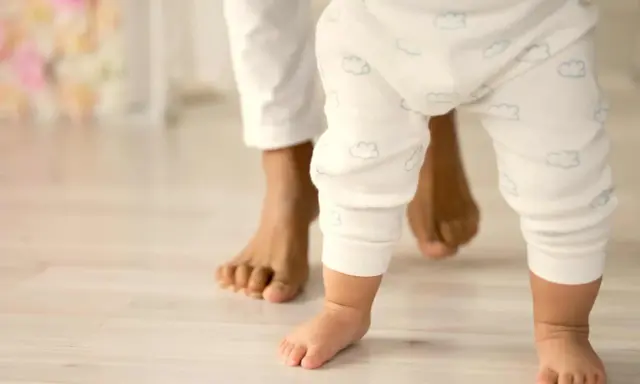 Un bebé da sus primeros pasos, guiado por un adulto. Un momento tierno que ilustra cómo educar a un bebé: con amor y apoyo.