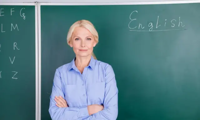 Nauczycielka angielskiego z uśmiechem, stojąca przed tablicą z napisem "English". Marzy o wcześniejszej emeryturze dla nauczycieli.