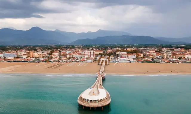 Un molo si estende su un mare azzurro, con una spiaggia dorata e una città costiera sullo sfondo. La Toscana offre un mare bello e panorami indimenticabili.