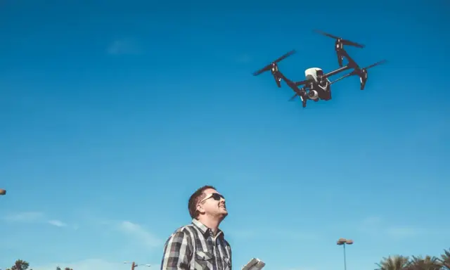 Ein **Drohnenpilot** steuert mit einer Fernbedienung einen Quadrocopter am blauen Himmel.