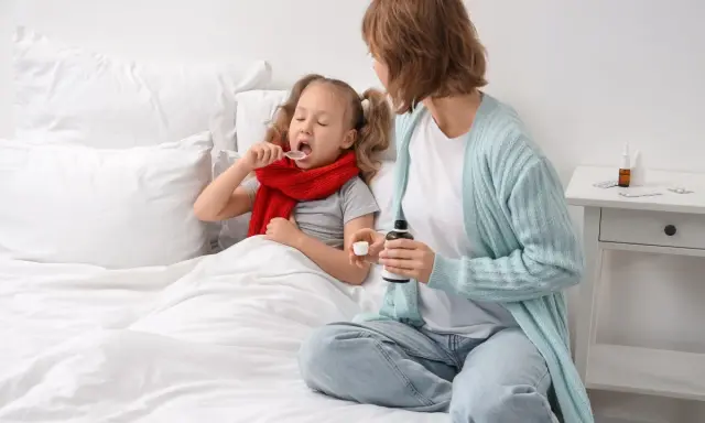 Bambina malata a letto prende sciroppo, mentre la mamma tiene il flacone. Forse per febbre o tosse, un mix di brufen e tachipirina insieme per farla stare meglio.