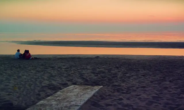 Alba a Rimini: una coppia seduta sulla sabbia guarda il mare calmo e il cielo colorato.