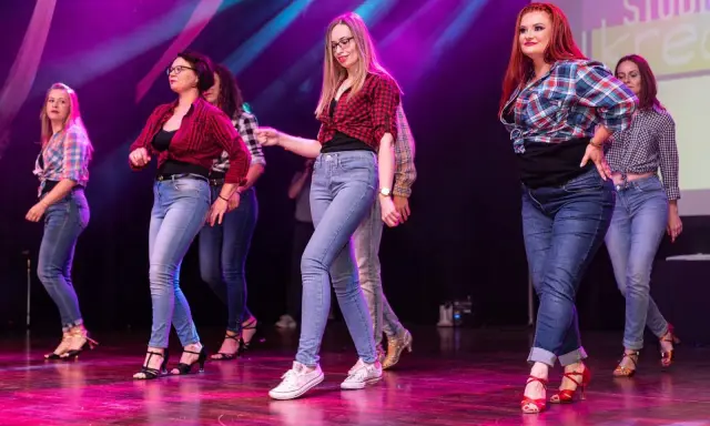Grupa kobiet w kratę i jeansach tańczy na scenie. Występ latino solo dla początkujących.