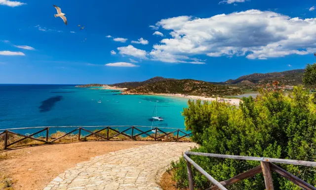 Vista panoramica di una baia con acque turchesi, spiagge affollate e montagne verdi. Un gabbiano vola nel cielo azzurro sopra Quartu e dintorni.
