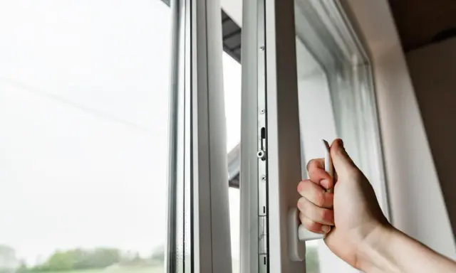 Mano girando manija de ventana blanca, asegurando la permeabilidad al aire clase 4. Ventana abierta con vista al exterior.