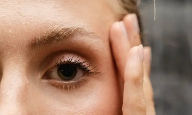 Un œil attentif, cils longs, et le début du repousse sourcils après chimio. Une main effleure le visage.