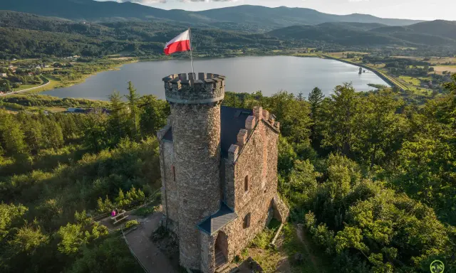 Kamienna wieża z polską flagą na tle jeziora i gór. Wspaniałe **cieplice atrakcje** dla miłośników historii i przyrody.