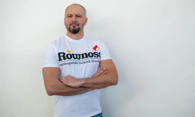 Mężczyzna w koszulce z napisem "Równość Ogólnopolski związek zawodowy" stoi z rękami skrzyżowanymi na piersi.