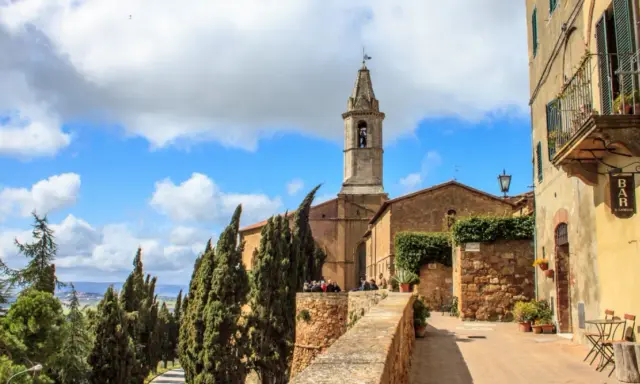 Un borgo nei comuni Val d'Orcia: un campanile svetta tra cipressi e case antiche, con un bar all'aperto.