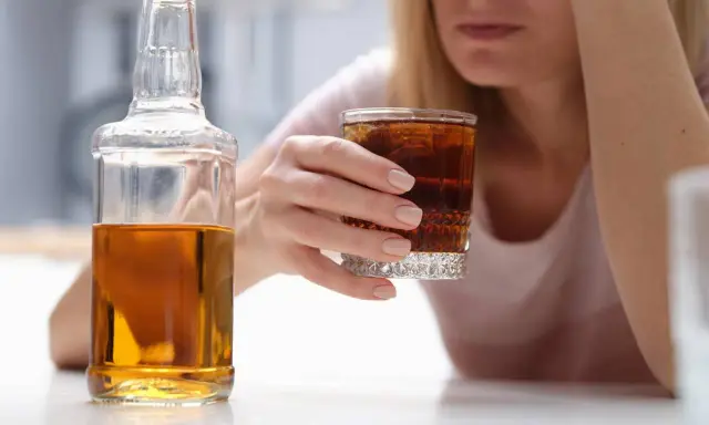 Kobieta z kieliszkiem drinka i butelką alkoholu. W tle widać jej twarz, która wyraża smutek. To może być wszywka alkoholowa.