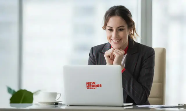 Uśmiechnięta kobieta przy laptopie z logo "New Heroes Academy" odkrywa, jak się przebranżowić.