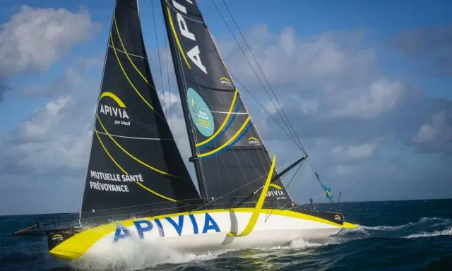 Le voilier Apivia, skippé par Alex Thomson, fend les vagues sous un ciel bleu et nuageux.